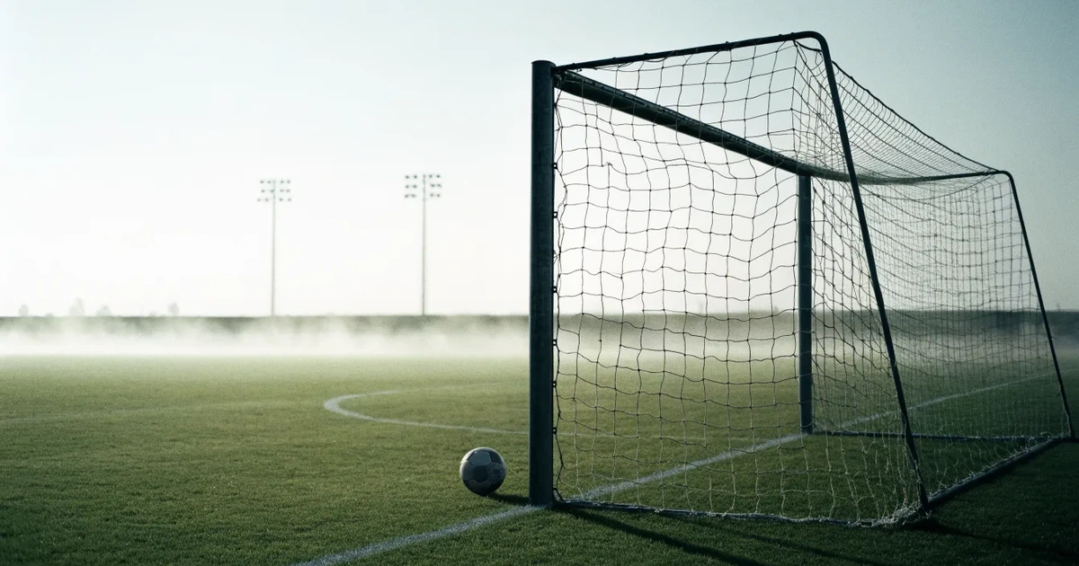 Ein Fußballtor mit gespanntem Netz steht auf einem leeren Rasenplatz, davor ein einzelner Ball, Morgendunst liegt über dem Feld.