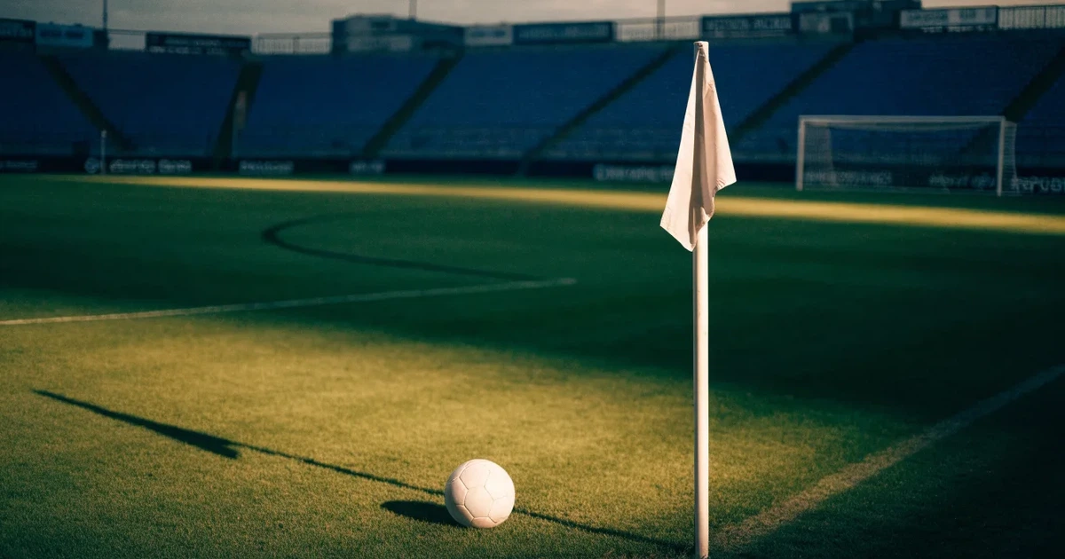 Blick durch ein Stadiontor auf einen grünen Rasen im Abendlicht, ein Ball liegt an der Eckfahne, die Ränge sind leer.