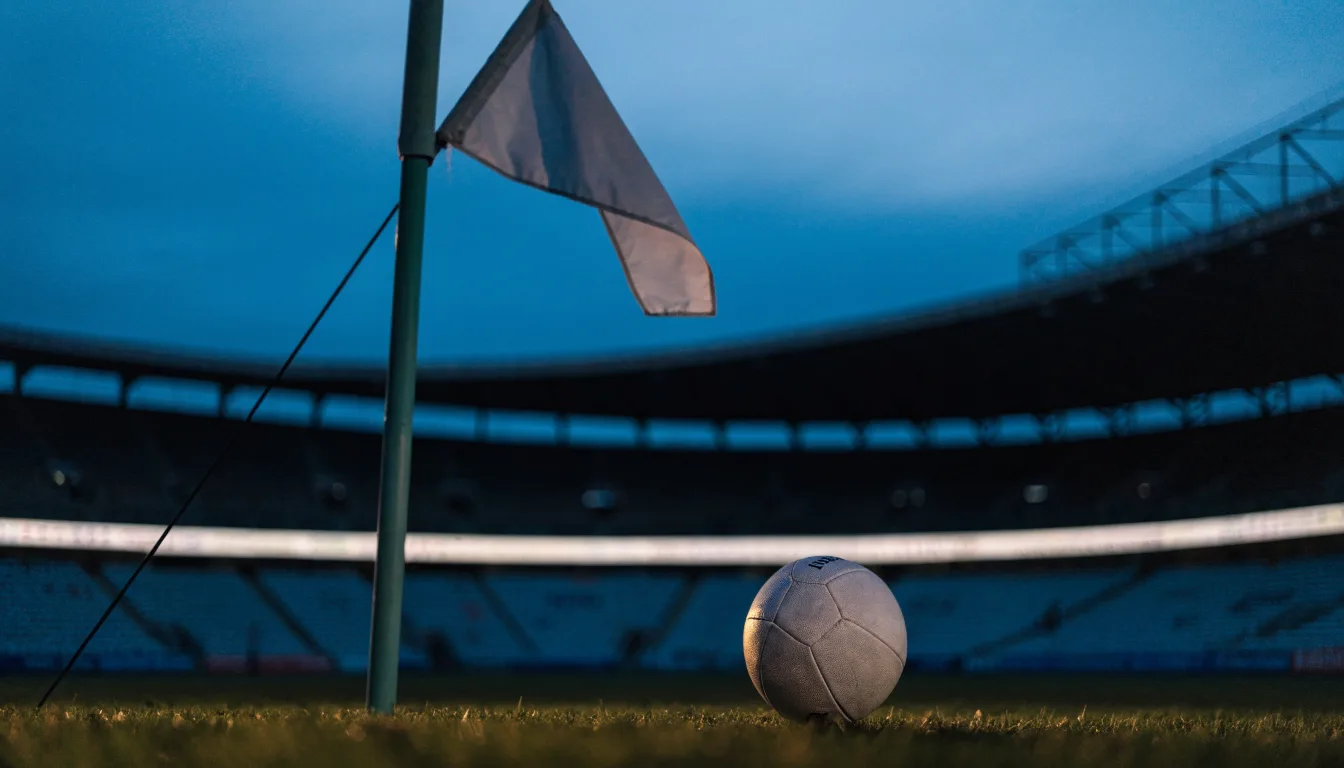 Ein Fußball liegt neben einem Eckfahnen-Pfosten auf einem Rasenplatz, im Hintergrund ein leeres Stadion in der Dämmerung.