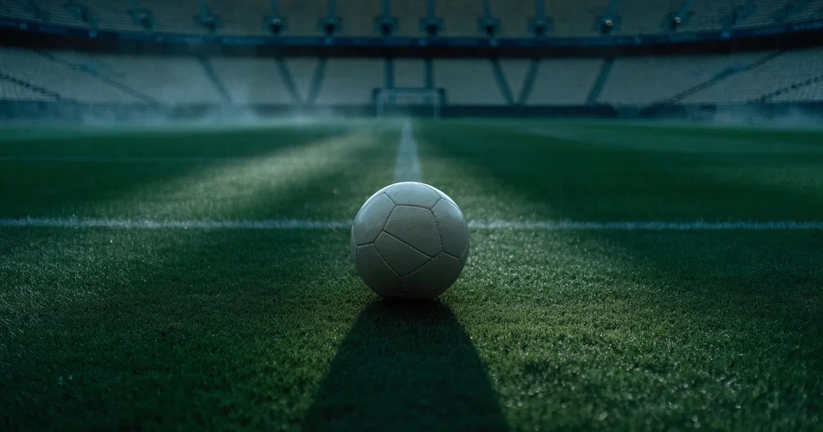 Ein Fußball liegt auf dem Mittelkreis eines Stadionrasens am frühen Morgen, im Hintergrund leere Ränge ohne erkennbare Beschriftung.