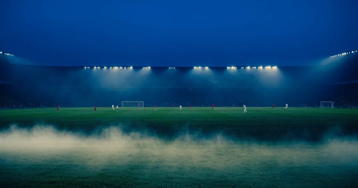 Ein Stadion unter Flutlicht, aus großer Entfernung über eine dunkle Wiese fotografiert, am Himmel zieht Nebel auf.