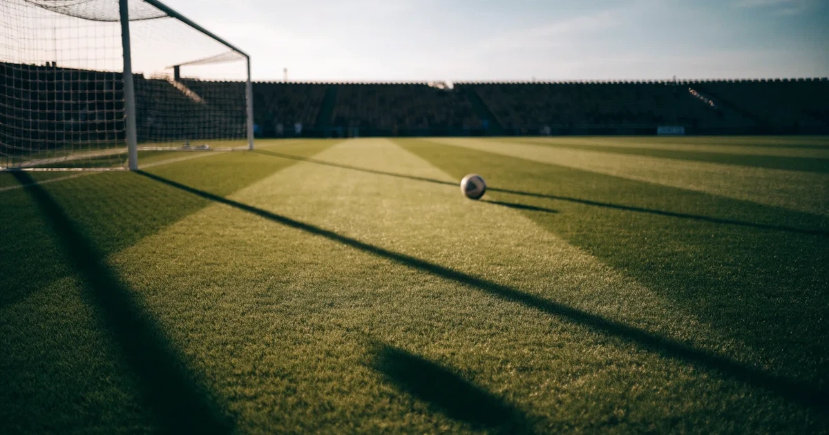 Ein Fußballplatz mit frisch gemähtem Rasen im Abendlicht, ein Ball liegt im Strafraum, im Hintergrund unscharf eine leere Tribüne.