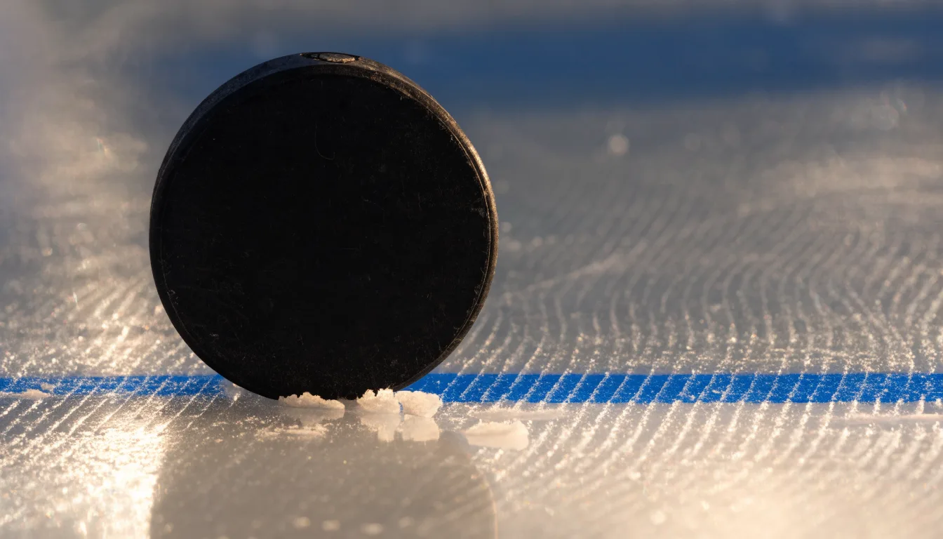 Ein schwarzer Puck liegt auf frisch aufbereitetem Eis neben der blauen Linie, Lichtreflexe auf der Fläche.