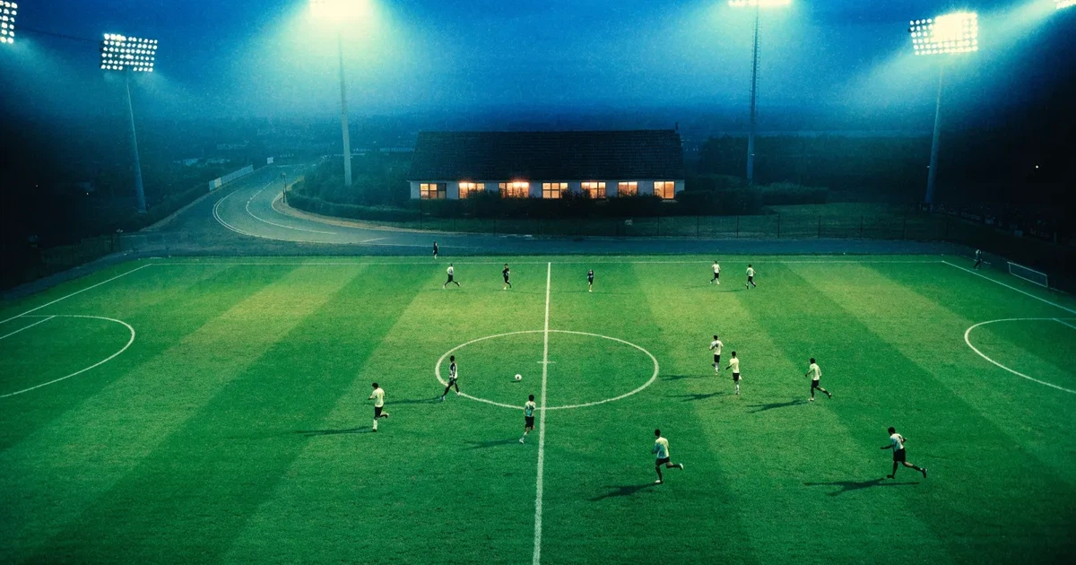 Ein Fußballstadion bei Flutlicht aus der Ferne gesehen, davor eine Straße und leuchtende Fenster eines Nebengebäudes.
