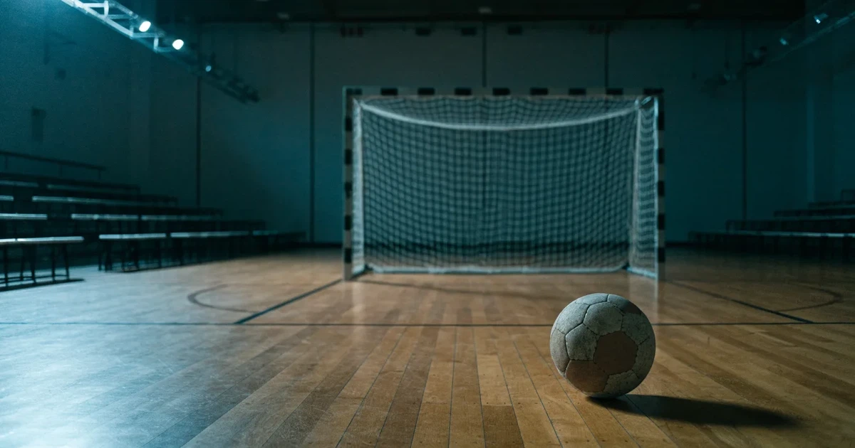 Ein Handball liegt auf dem Hallenboden vor einem Tor mit Netz, die leere Halle wird von Flutlicht beleuchtet.
