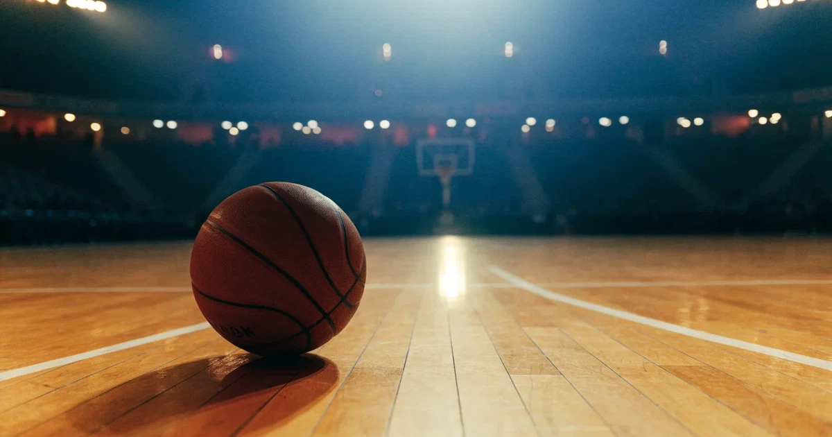 Ein Basketball liegt auf dem Parkettboden einer leeren Arena, Scheinwerfer werfen Licht auf die Mittellinie.