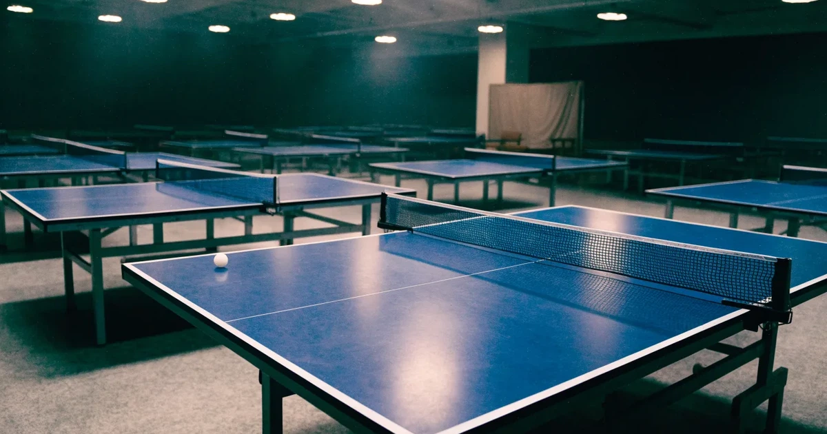 Eine Tischtennishalle aus erhöhter Perspektive mit mehreren blauen Tischen in Reihen unter gedämpftem Hallenlicht, ohne Personen.