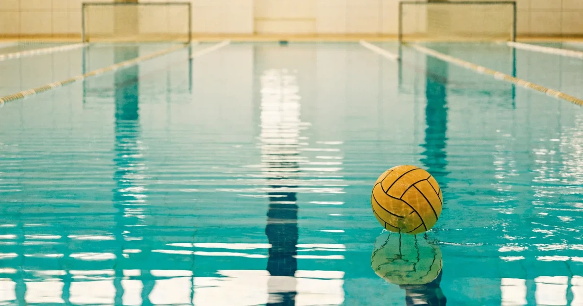 Ein Schwimmbecken mit türkisfarbenem Wasser und zwei schwimmenden Torrahmen am gegenüberliegenden Beckenrand, menschenleer unter Hallenlicht.