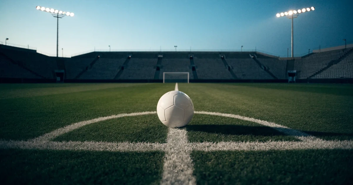 Ein Rasenplatz in der Dämmerung mit eingeschalteten Flutlichtmasten, ein Ball liegt am Anstoßpunkt, die Tribünen im Hintergrund sind unscharf.