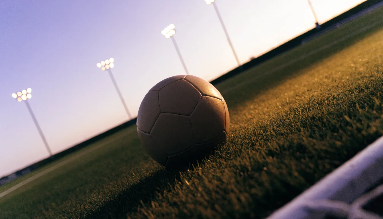Ein Fußball liegt im Gras eines leeren Platzes, im Hintergrund stehen Flutlichtmasten in der Dämmerung.