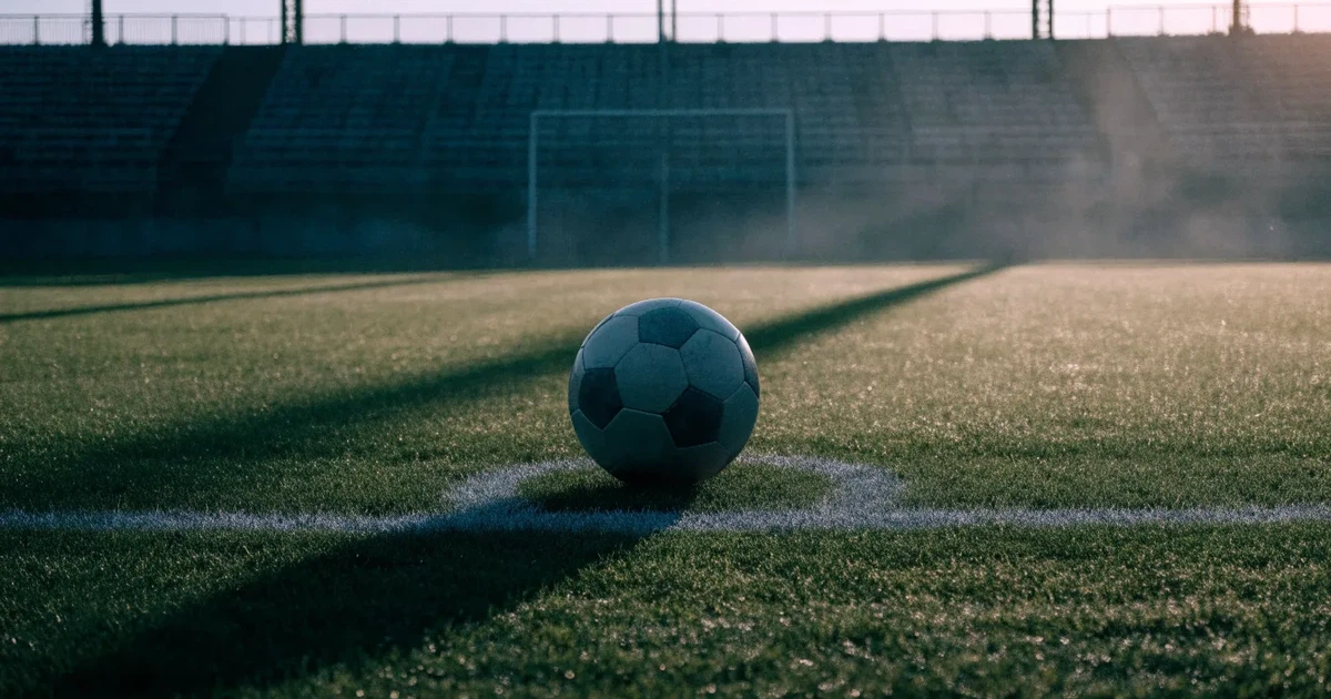 Ein Fußball liegt auf dem Anstoßpunkt eines Rasenplatzes, dahinter leere Tribünen im Licht des frühen Morgens.