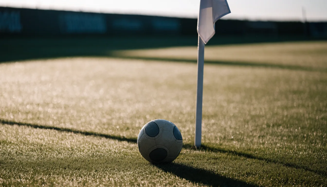 Ein Fußball liegt auf Rasen neben einer Eckfahne ohne Aufdruck auf einem leeren Platz im Morgenlicht.