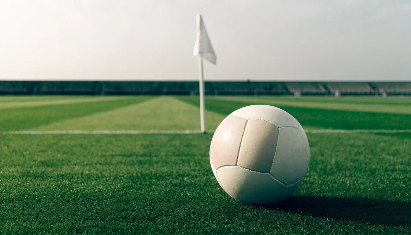 Ein Fußball liegt neben einer Eckfahne auf kurz gemähtem Rasen unter bewölktem Himmel.